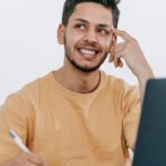 Future Strategies - Smiling young bearded Hispanic male entrepreneur thinking over new ideas for startup project and looking away dreamily while working at table with laptop and taking notes in notebook