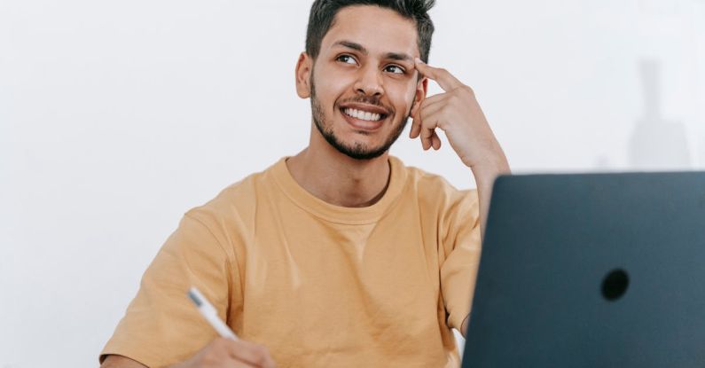 Future Strategies - Smiling young bearded Hispanic male entrepreneur thinking over new ideas for startup project and looking away dreamily while working at table with laptop and taking notes in notebook