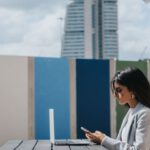Startup Building - Ethnic female executive watching smartphone at table with laptop outdoors