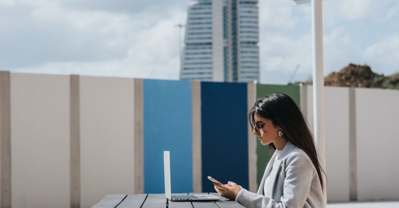 Startup Building - Ethnic female executive watching smartphone at table with laptop outdoors