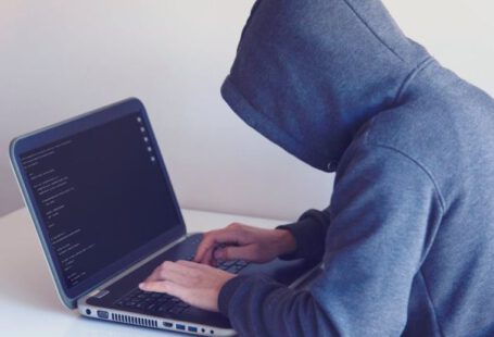 Fraud Protection - Side view of unrecognizable hacker in hoodie sitting at white table and working remotely on netbook in light room near wall