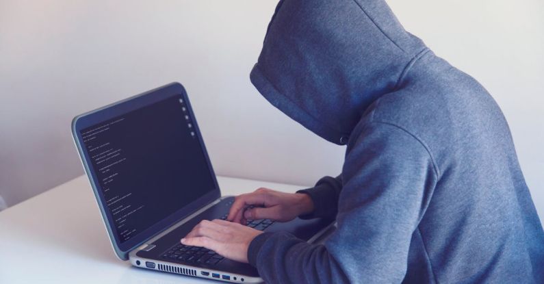 Fraud Protection - Side view of unrecognizable hacker in hoodie sitting at white table and working remotely on netbook in light room near wall